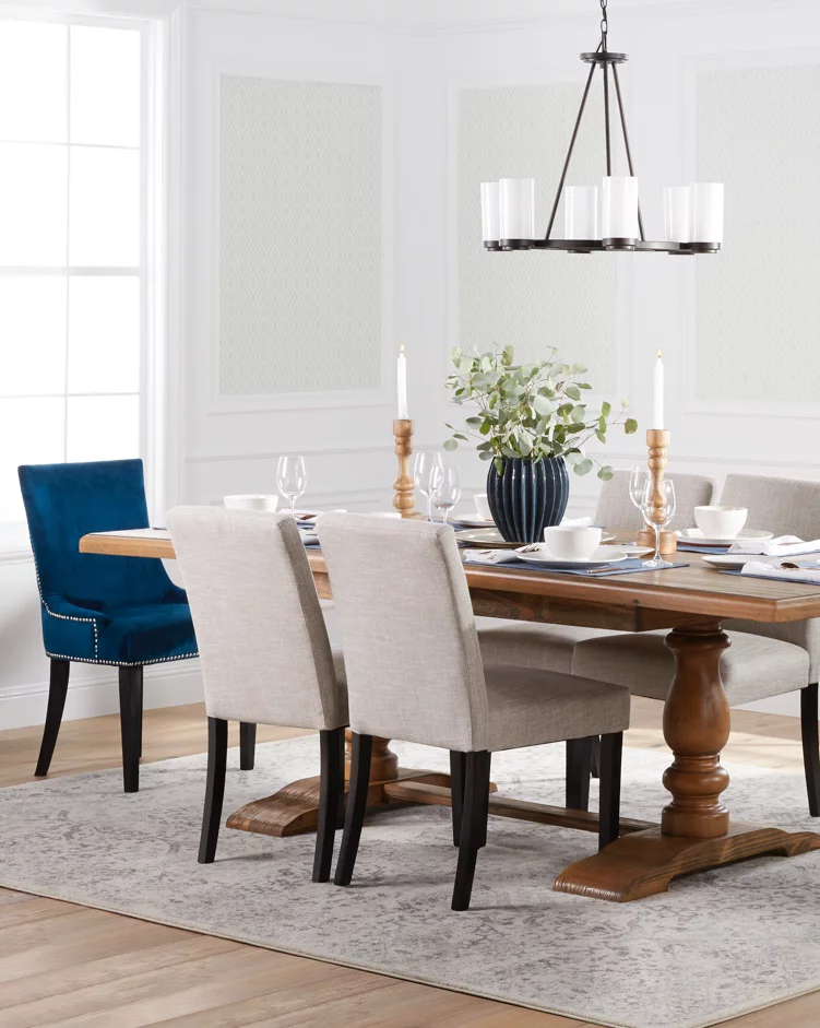 A transitional dining room with a beautiful wooden trestle table, upholstered seating, blue velvet end chairs, an industrial chandelier and a dining table area rug. Links to DX Fair Mall's dining room furniture and decor